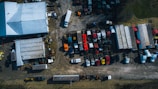 An overhead view of a large yard featuring a collection of trucks and trailers parked in rows. A warehouse with a white roof is positioned on the left. The area has a mixed surface of concrete and gravel, and multiple vehicles are scattered across the lot in organized sections. Surrounding the yard is a strip of grass, with various shadows cast by the trucks and structures.
