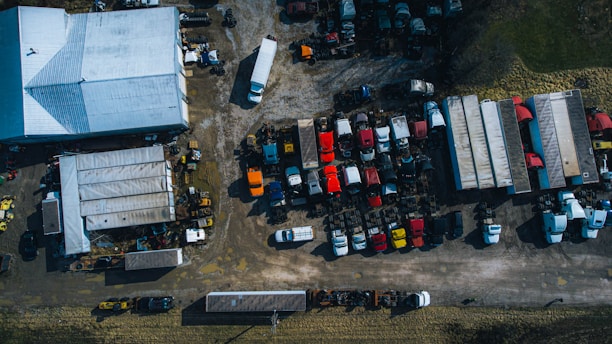 An overhead view of a large yard featuring a collection of trucks and trailers parked in rows. A warehouse with a white roof is positioned on the left. The area has a mixed surface of concrete and gravel, and multiple vehicles are scattered across the lot in organized sections. Surrounding the yard is a strip of grass, with various shadows cast by the trucks and structures.