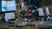 An overhead view of a large yard featuring a collection of trucks and trailers parked in rows. A warehouse with a white roof is positioned on the left. The area has a mixed surface of concrete and gravel, and multiple vehicles are scattered across the lot in organized sections. Surrounding the yard is a strip of grass, with various shadows cast by the trucks and structures.