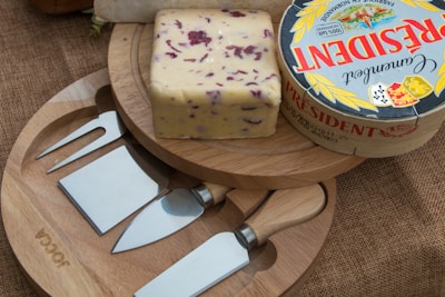A block of cheese with cranberries sits on a wooden board alongside a round box of President Camembert cheese. There are three types of cheese knives arranged neatly on a round wooden board beneath the cheeses. The background surface is textured and brown, resembling burlap.