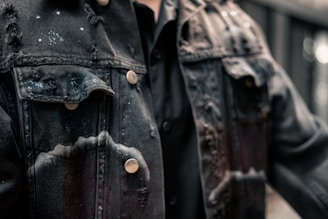 A close-up view of a black denim jacket featuring a distressed texture with several frayed edges and bleached spots. The material has a rugged appearance, complemented by metallic buttons. It appears to be worn over a black shirt.