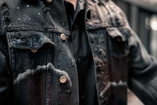 A close-up view of a black denim jacket featuring a distressed texture with several frayed edges and bleached spots. The material has a rugged appearance, complemented by metallic buttons. It appears to be worn over a black shirt.