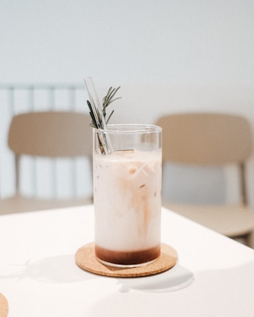 A clear glass filled with iced coffee featuring a layered appearance, with milk and coffee separated. A clear straw and a sprig of rosemary are added for garnish. The glass is placed on a round cork coaster on a light-colored table. The background includes blurred, light beige chairs and a minimalistic setting.