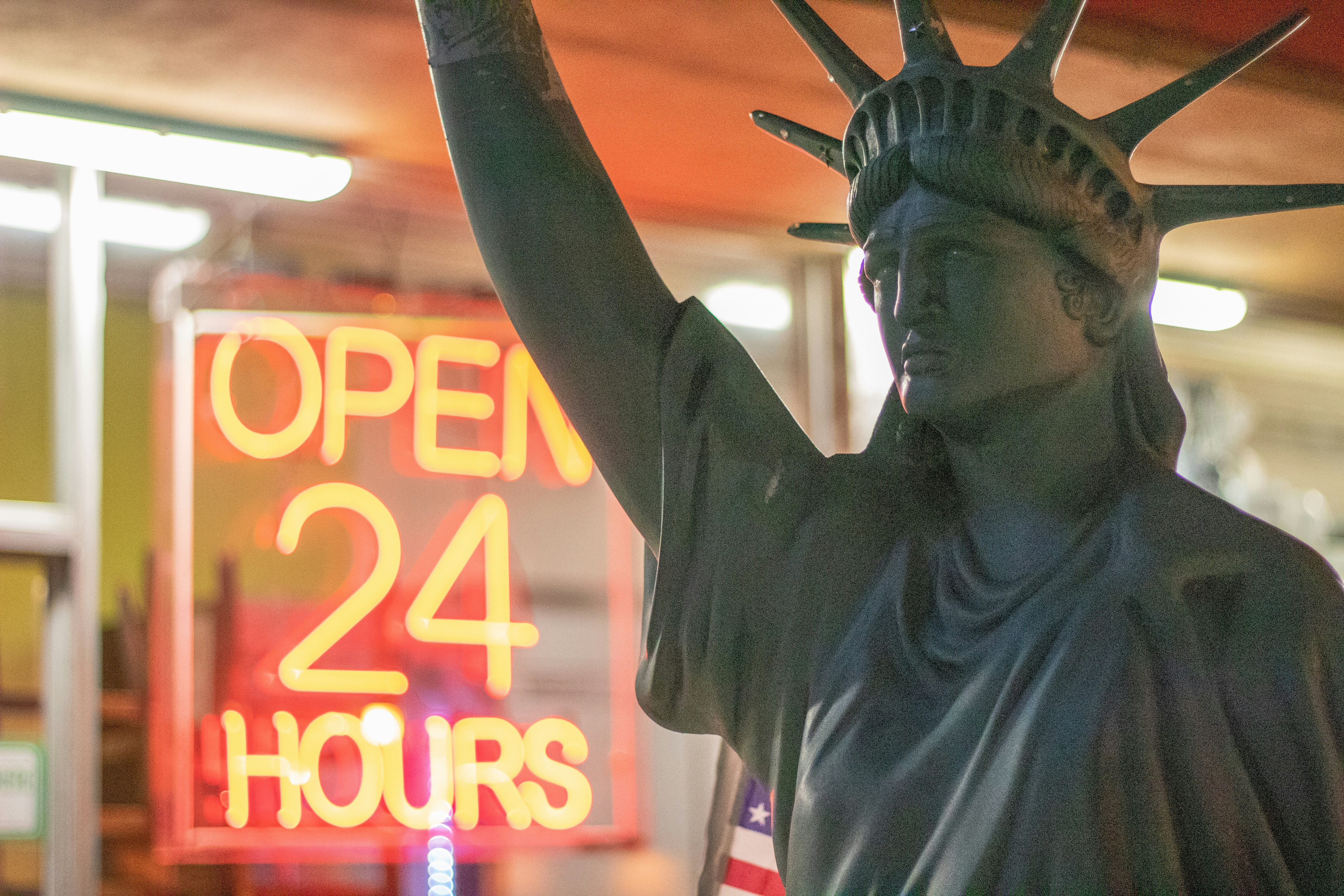 man in white crew neck t-shirt, Statue of Liberty at a taco shop holding a burrito with Open 24 Hours sign. 