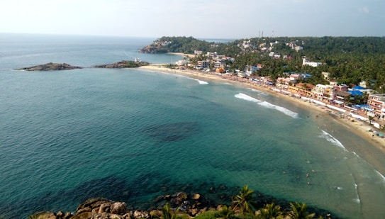 Aerial view of a coastal area with a curved sandy beach lined with numerous buildings, primarily colorful hotels and houses. The right side features lush greenery with coconut trees. The calm blue ocean stretches out, meeting the shoreline gently, while a rocky outcrop extends into the sea.