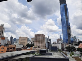 A cityscape featuring a modern skyline with a prominent skyscraper clad in reflective blue glass. Older, lower-rise buildings in beige and orange hues are in the foreground, set against a backdrop of additional skyscrapers under a partly cloudy sky.