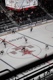 An ice hockey game is being played in a large arena. Two teams, one in red jerseys and the other in white, are on the ice with players and referees. The arena has digital screens showing game details above the spectators who are seated in the stands. The view is from an elevated position, capturing the center ice area and the surrounding ring with branded advertisements.