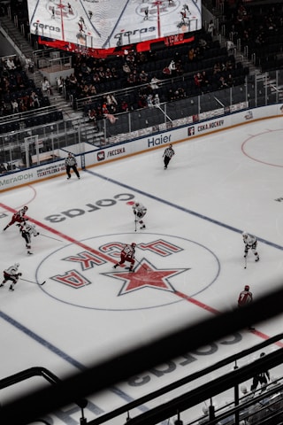 An ice hockey game is being played in a large arena. Two teams, one in red jerseys and the other in white, are on the ice with players and referees. The arena has digital screens showing game details above the spectators who are seated in the stands. The view is from an elevated position, capturing the center ice area and the surrounding ring with branded advertisements.