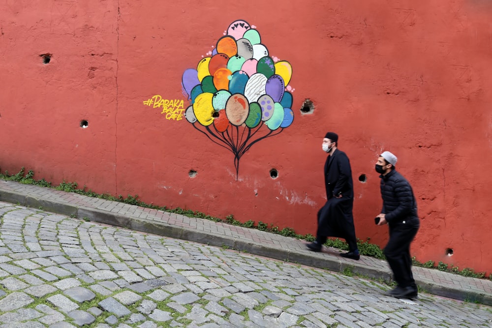 people walking on sidewalk near red wall with yellow blue and red heart shaped wall art