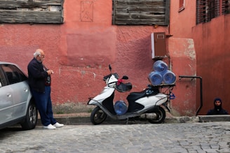 An elderly man stands next to a parked scooter loaded with large water bottles, which is situated beside a car on a cobblestone street. The background features a worn pink wall with wooden and brick sections. A person in dark clothing is partially visible at the bottom of a staircase leading underground.