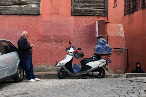 An elderly man stands next to a parked scooter loaded with large water bottles, which is situated beside a car on a cobblestone street. The background features a worn pink wall with wooden and brick sections. A person in dark clothing is partially visible at the bottom of a staircase leading underground.