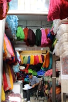 A small shop is filled with colorful bundles of yarn hanging from the ceiling and walls. A person sits on a chair, reviewing some documents at a small desk in the corner. The shop is vibrant with various colors of yarn including red, green, blue, yellow, and pink, creating a vibrant and textured environment.