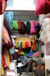A small shop is filled with colorful bundles of yarn hanging from the ceiling and walls. A person sits on a chair, reviewing some documents at a small desk in the corner. The shop is vibrant with various colors of yarn including red, green, blue, yellow, and pink, creating a vibrant and textured environment.