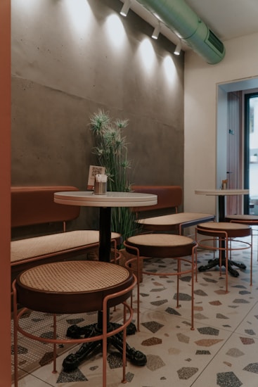 A cozy corner of a caf&eacute; with retro-style tables and chairs. The seating is made of wicker and metal frames, set against a textured wall with subtle lighting fixtures near the ceiling. Decorative plants add a touch of greenery, and a QR code stand is placed on the table. Light streams in softly from a nearby window, illuminating a terrazzo floor.