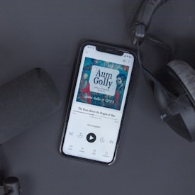 A smartphone displaying an audiobook app screen with a cover titled 'Aum Golly' and subtitle 'The Poem About the Origins of Man.' The phone is on a dark surface with a pair of headphones and a microphone visible nearby. The image composition suggests a podcast or audiobook listening setup.