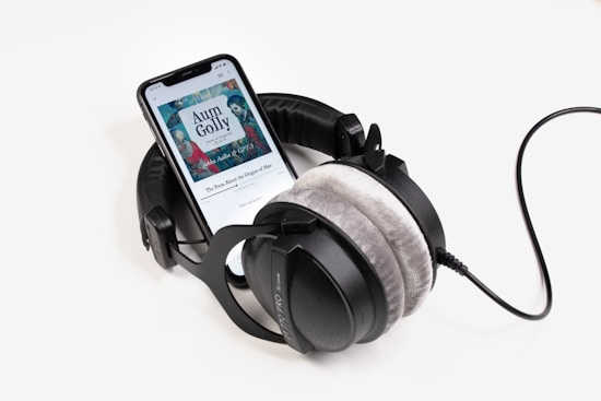 A pair of large over-ear headphones is placed around a smartphone displaying an audiobook titled 'Aum Golly'. The headphones have plush gray ear cushions and are connected with a thick black wire.