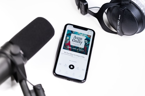 A smartphone displays an audiobook app with the cover art for 'Aum Golly' by Hokusai on its screen. Nearby, a large black microphone and a set of black studio headphones are placed on a white surface, suggesting an audio recording or podcast setup.
