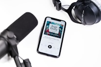 A smartphone displays an audiobook app with the cover art for 'Aum Golly' by Hokusai on its screen. Nearby, a large black microphone and a set of black studio headphones are placed on a white surface, suggesting an audio recording or podcast setup.