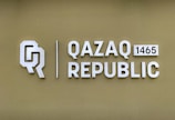 The image features a logo with the letters 'Q' and 'R' next to the text 'QAZAQ REPUBLIC' and the number '1465'. The background is a muted olive green color, and the text and logo are in a light, metallic shade.