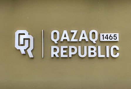 The image features a logo with the letters 'Q' and 'R' next to the text 'QAZAQ REPUBLIC' and the number '1465'. The background is a muted olive green color, and the text and logo are in a light, metallic shade.