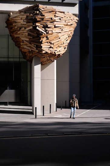 A large architectural installation made of wooden planks is attached to the exterior of a modern building. It resembles a giant, abstract nest or beehive. A person wearing a brown jacket and jeans is walking on the sidewalk next to the structure.