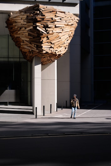 A large architectural installation made of wooden planks is attached to the exterior of a modern building. It resembles a giant, abstract nest or beehive. A person wearing a brown jacket and jeans is walking on the sidewalk next to the structure.