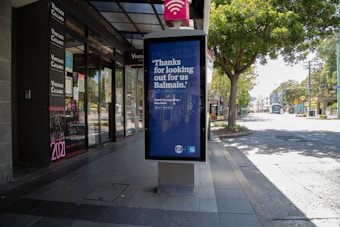 A quiet urban street scene with a row of shops on the left side, featuring a prominent storefront for Vintage Cellars. In the foreground, a large advertisement board with a message thanking the community is strategically positioned. The street is lined with trees and appears serene with no visible pedestrians or vehicles.