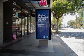 A quiet urban street scene with a row of shops on the left side, featuring a prominent storefront for Vintage Cellars. In the foreground, a large advertisement board with a message thanking the community is strategically positioned. The street is lined with trees and appears serene with no visible pedestrians or vehicles.