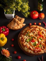 A freshly baked pizza topped with colorful ingredients such as peppers, olives, and cheese is placed on a wooden platter. The setting includes a variety of fresh ingredients like whole tomatoes, red and yellow bell peppers, and a bottle of olive oil. Freshly baked bread is placed nearby along with decorative flowers and leafy plants, creating a vibrant and inviting culinary scene.