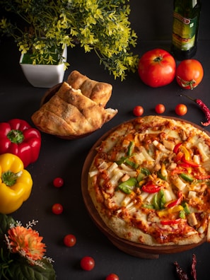 A freshly baked pizza topped with colorful ingredients such as peppers, olives, and cheese is placed on a wooden platter. The setting includes a variety of fresh ingredients like whole tomatoes, red and yellow bell peppers, and a bottle of olive oil. Freshly baked bread is placed nearby along with decorative flowers and leafy plants, creating a vibrant and inviting culinary scene.