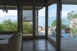 A serene indoor space with wooden features and large windows overlooks a beautiful outdoor view of a calm sea with distant islands. There is a sofa with cushions inside, and outside, there are lounge chairs next to a small pool surrounded by greenery.