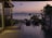 A serene and tranquil outdoor scene features an infinity pool overlooking a calm sea, set against a backdrop of distant hills and a colorful sunset sky. There are two lounge chairs positioned by the pool, surrounded by natural elements such as wooden structures, lush greenery, and bamboo fencing.