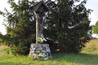 A wooden memorial structure with a roof stands on a stone base. It is surrounded by green grass and large evergreen trees. White flowers are placed at the base of the structure, suggesting a tribute or memorial.