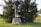 A wooden memorial structure with a roof stands on a stone base. It is surrounded by green grass and large evergreen trees. White flowers are placed at the base of the structure, suggesting a tribute or memorial.