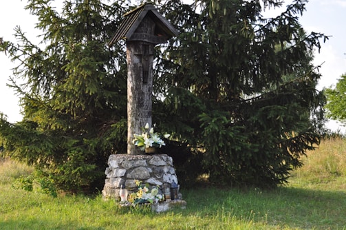 A wooden memorial structure with a roof stands on a stone base. It is surrounded by green grass and large evergreen trees. White flowers are placed at the base of the structure, suggesting a tribute or memorial.