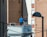Two people are standing on a rooftop next to a prominent, curved sign displaying a dental clinic's services. One person is wearing a blue top, and the other is in a dark outfit with a red hat. The background features tall brick buildings, indicating an urban setting. A lamp post and several wires are visible in the foreground.