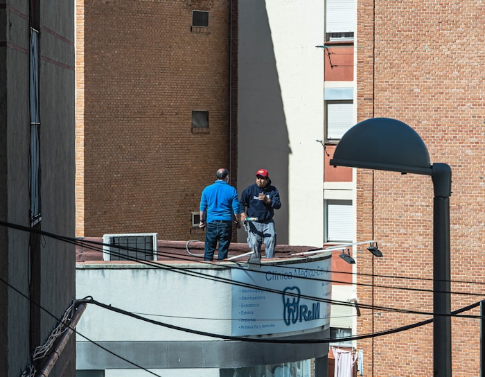 Two people are standing on a rooftop next to a prominent, curved sign displaying a dental clinic's services. One person is wearing a blue top, and the other is in a dark outfit with a red hat. The background features tall brick buildings, indicating an urban setting. A lamp post and several wires are visible in the foreground.