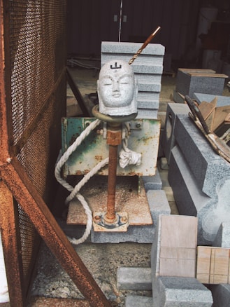 A sculpture of a head is attached to a metal structure with a metal rod protruding from the top. The scene is surrounded by stacked stone slabs and blocks. The area appears to be an industrial or workshop setting, with visible rust on the metal and various construction or sculpting materials around.