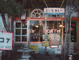 A cozy cafe entrance with vintage signage and an eclectic mix of decorations. The building features arched windows and colorful signs advertising various menu items in Japanese and English. A stone lantern is situated at the front, surrounded by decorative rocks and a small tree, adding to the peaceful ambiance.