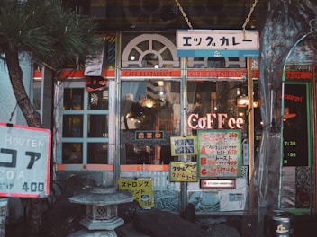 A cozy cafe entrance with vintage signage and an eclectic mix of decorations. The building features arched windows and colorful signs advertising various menu items in Japanese and English. A stone lantern is situated at the front, surrounded by decorative rocks and a small tree, adding to the peaceful ambiance.