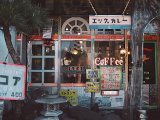 A cozy cafe entrance with vintage signage and an eclectic mix of decorations. The building features arched windows and colorful signs advertising various menu items in Japanese and English. A stone lantern is situated at the front, surrounded by decorative rocks and a small tree, adding to the peaceful ambiance.