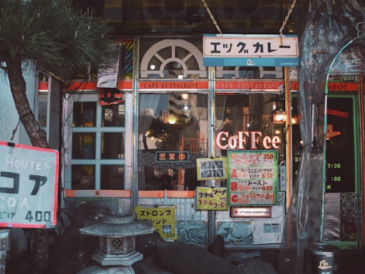 A cozy cafe entrance with vintage signage and an eclectic mix of decorations. The building features arched windows and colorful signs advertising various menu items in Japanese and English. A stone lantern is situated at the front, surrounded by decorative rocks and a small tree, adding to the peaceful ambiance.
