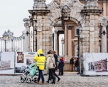 A historic stone archway with intricate carvings serves as the gateway to a building with classical architecture. People in winter clothing, including a person in a bright yellow reflective jacket, are walking and pushing a stroller beneath the arch. In the background, several individuals are observing historical photographs displayed on large boards.