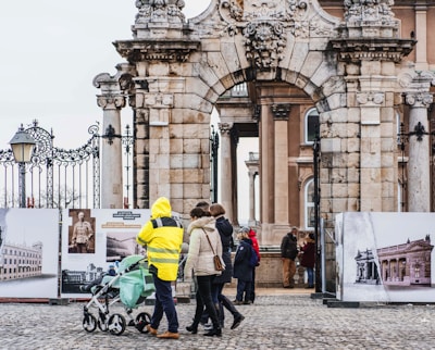 A historic stone archway with intricate carvings serves as the gateway to a building with classical architecture. People in winter clothing, including a person in a bright yellow reflective jacket, are walking and pushing a stroller beneath the arch. In the background, several individuals are observing historical photographs displayed on large boards.