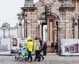 A historic stone archway with intricate carvings serves as the gateway to a building with classical architecture. People in winter clothing, including a person in a bright yellow reflective jacket, are walking and pushing a stroller beneath the arch. In the background, several individuals are observing historical photographs displayed on large boards.
