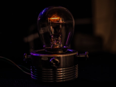 A vintage light bulb is encased in a metallic holder with screws, set against a dark background. The bulb is partially illuminated, casting a warm, dim glow. The overall ambiance is one of warmth and subtle sophistication.