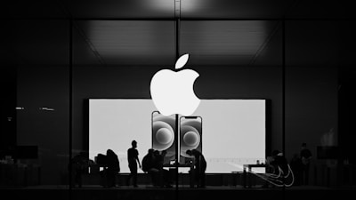 Silhouettes of people inside an Apple store appear against a backdrop of a large Apple logo and images of smartphones. The scene takes place during night or evening, creating a contrast between the bright interior and the dark exterior.