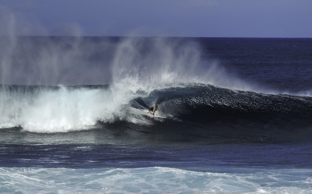 A surfer is captured riding a large wave, with white spray and mist forming as the wave curls over. The ocean appears deep and powerful, with contrasting dark and light hues. The scene conveys a sense of motion and energy.