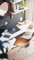 A stylish modern workspace features a white desk against a textured dark wall, with two open shelves holding books, decorative items, and a small plant. A sleek chair with a wooden seat and metal legs is positioned at the desk. There is a round mirror on the wall and a closed laptop on the desk. Natural light enters from the right, creating a warm and inviting atmosphere.