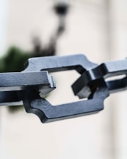 A close-up of a metal chain link against a blurred background. The focus is sharp on the chain, showing its geometric shapes and rugged texture. The background is light and softly out of focus, bringing attention to the metallic object.